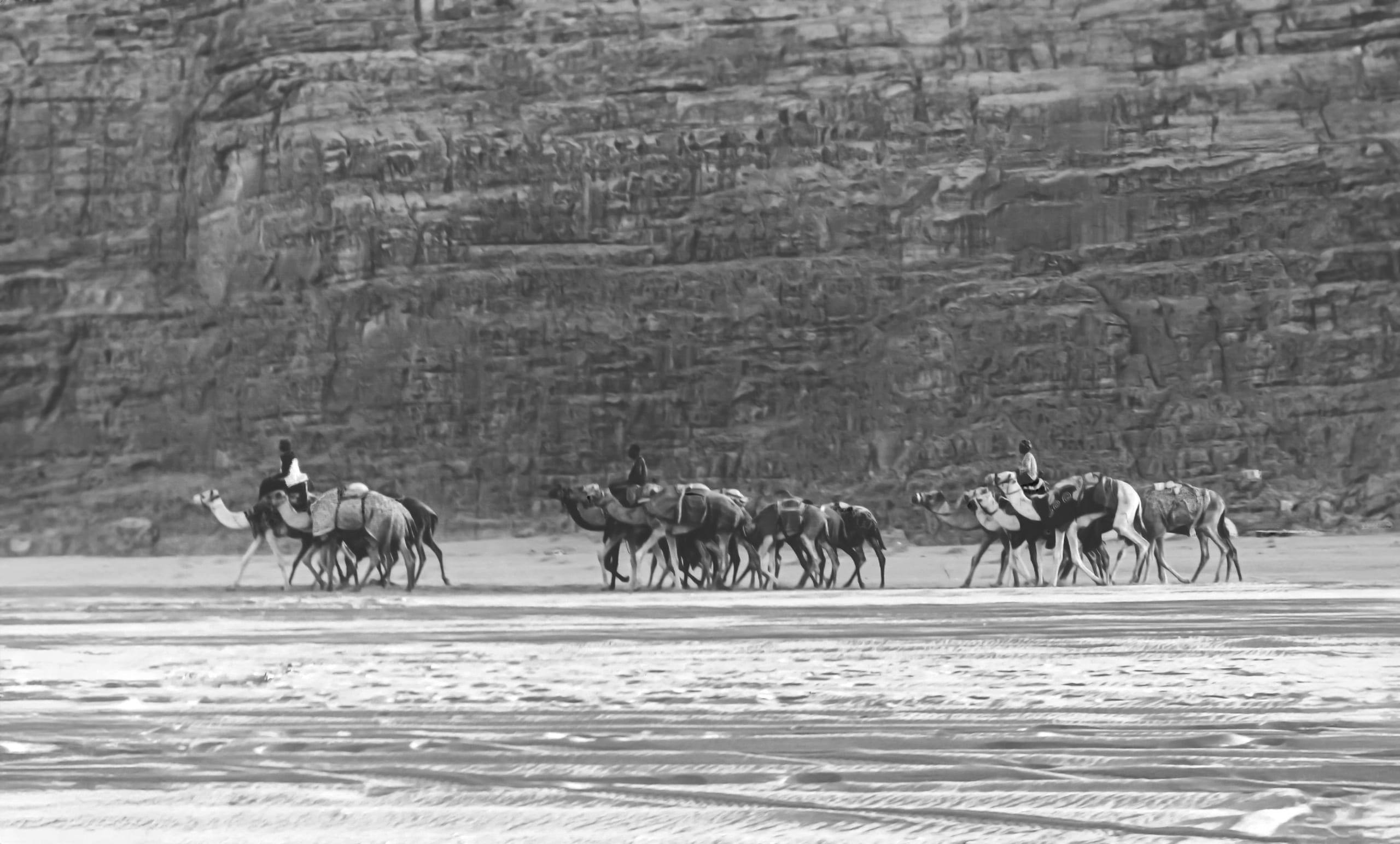 Monochrome photograph of a camel caravan with riders traveling across a wide, flat desert floor past a massive, textured cliff face.