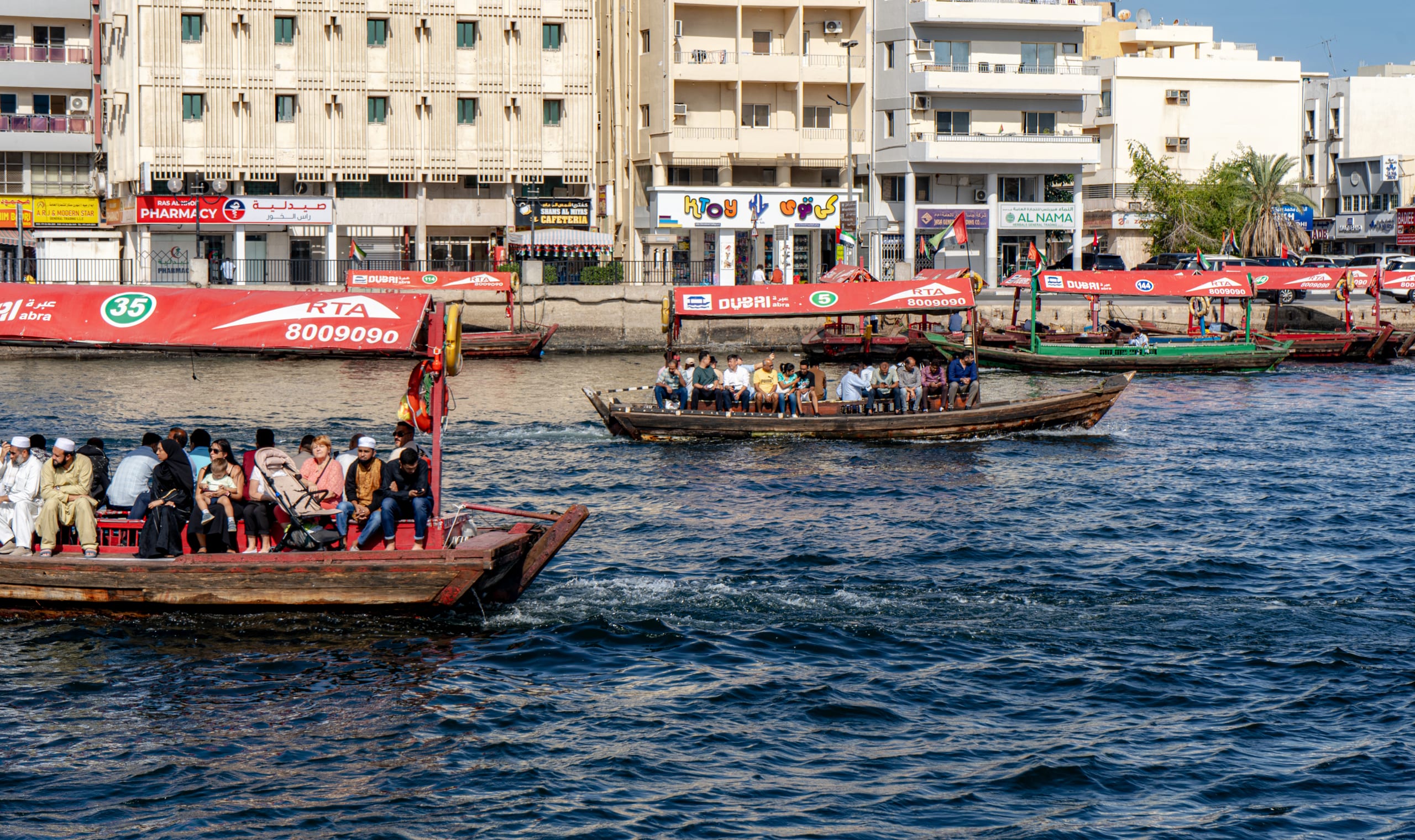 Multiple traditional wooden passenger boats filled with people crossing a blue water channel, with low-rise apartment and retail buildings lining the far bank under bright daylight.