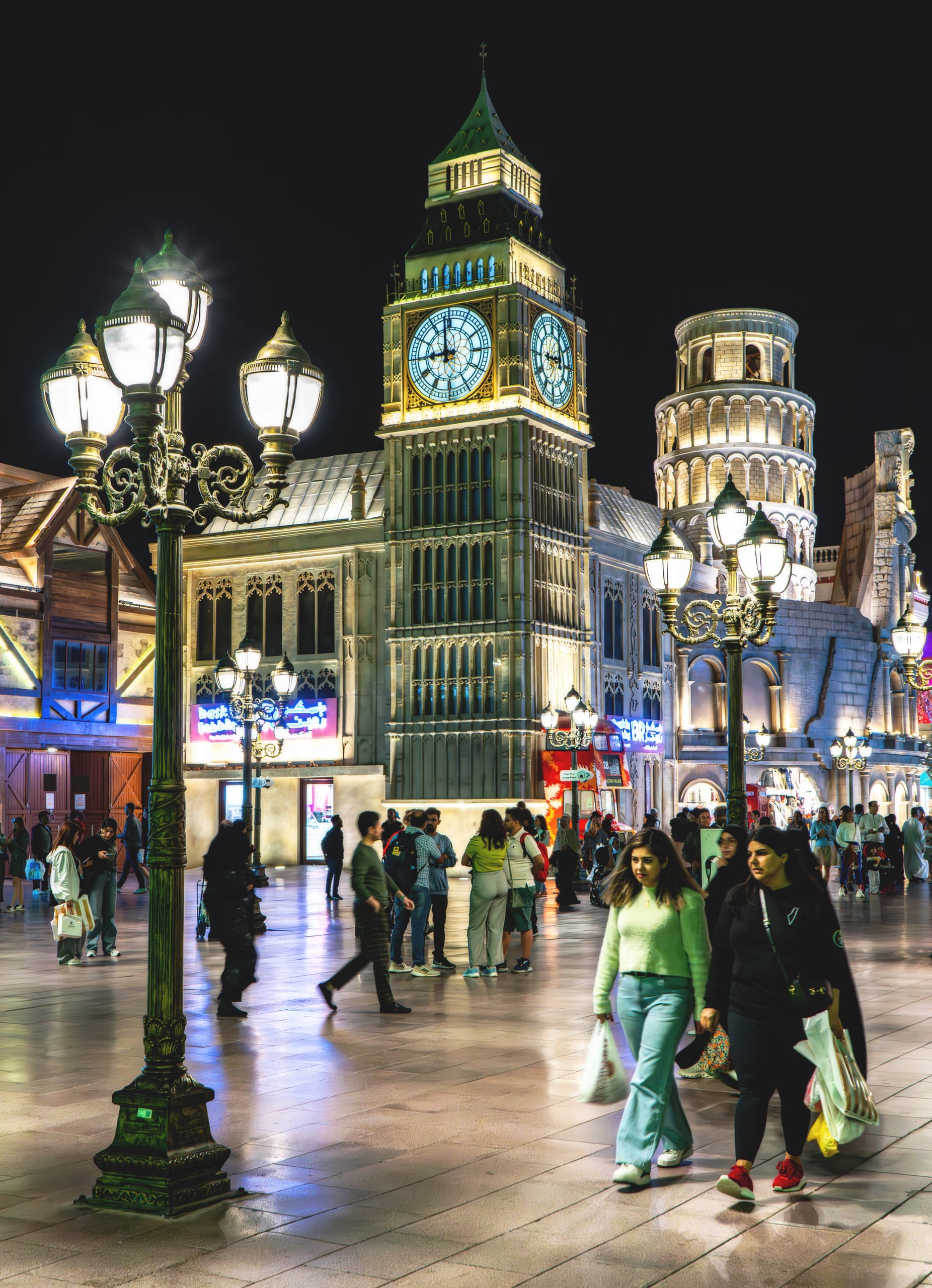 Pedestrians walking on a polished stone plaza at night beneath elaborate traditional streetlamps, with a lit clock tower and classical facades in the background.