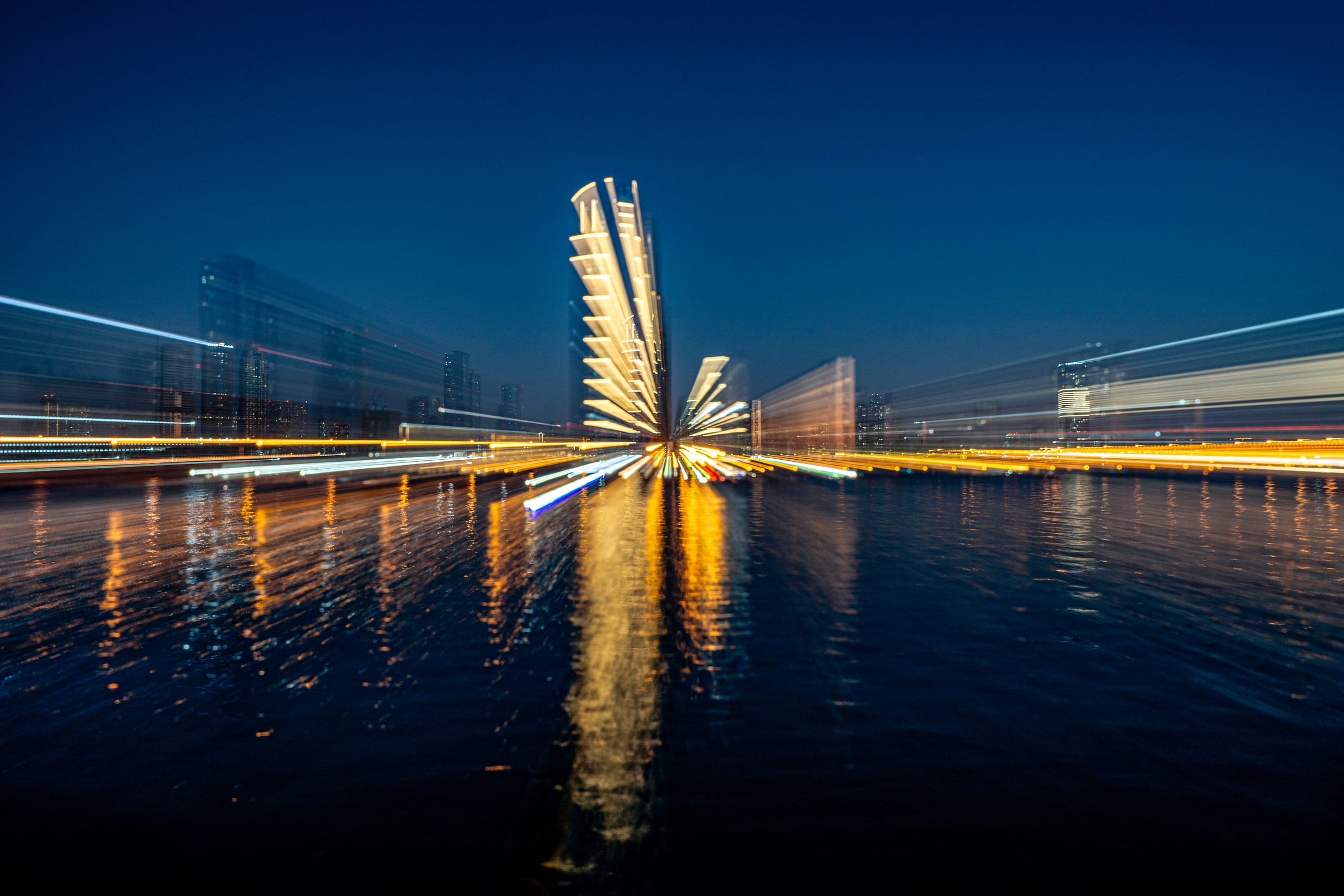 Long-exposure night photograph of a city skyline with radial zoom blur creating streaks of white and golden light radiating outward across dark water.