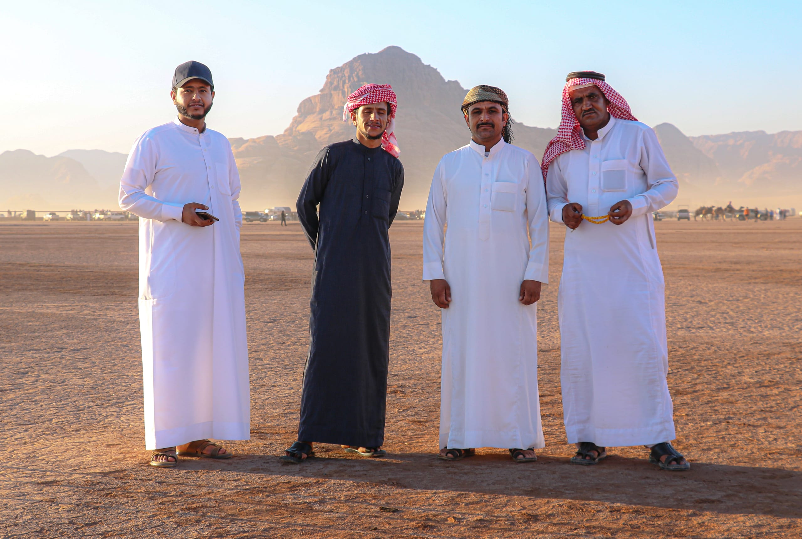 Four men in traditional clothing standing in a row in an open desert landscape with mountains in the background under hazy light.