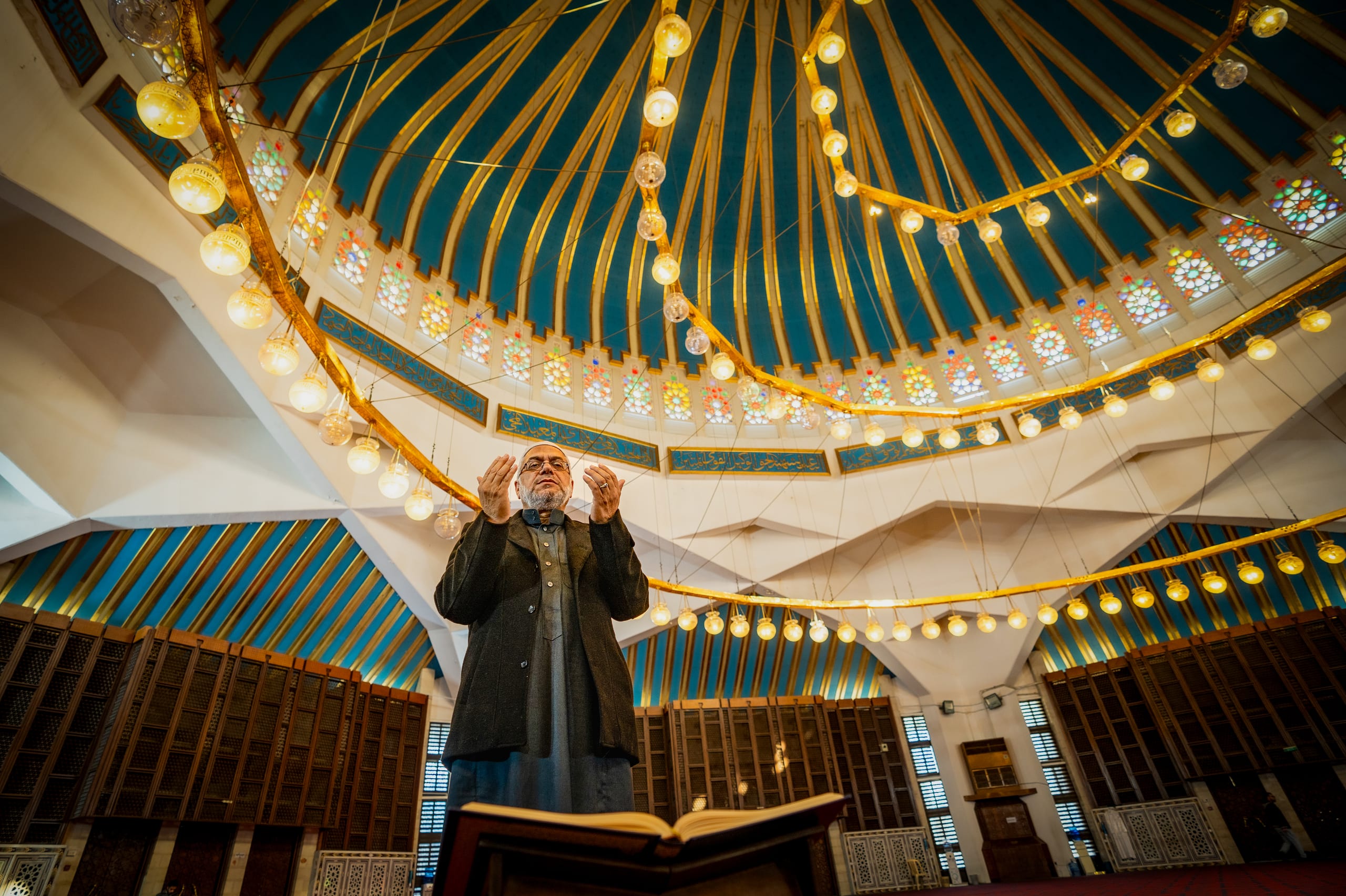 An elderly man standing inside a large hall with arms raised in prayer beneath a high domed ceiling featuring radial ribs and circular illuminated chandeliers.