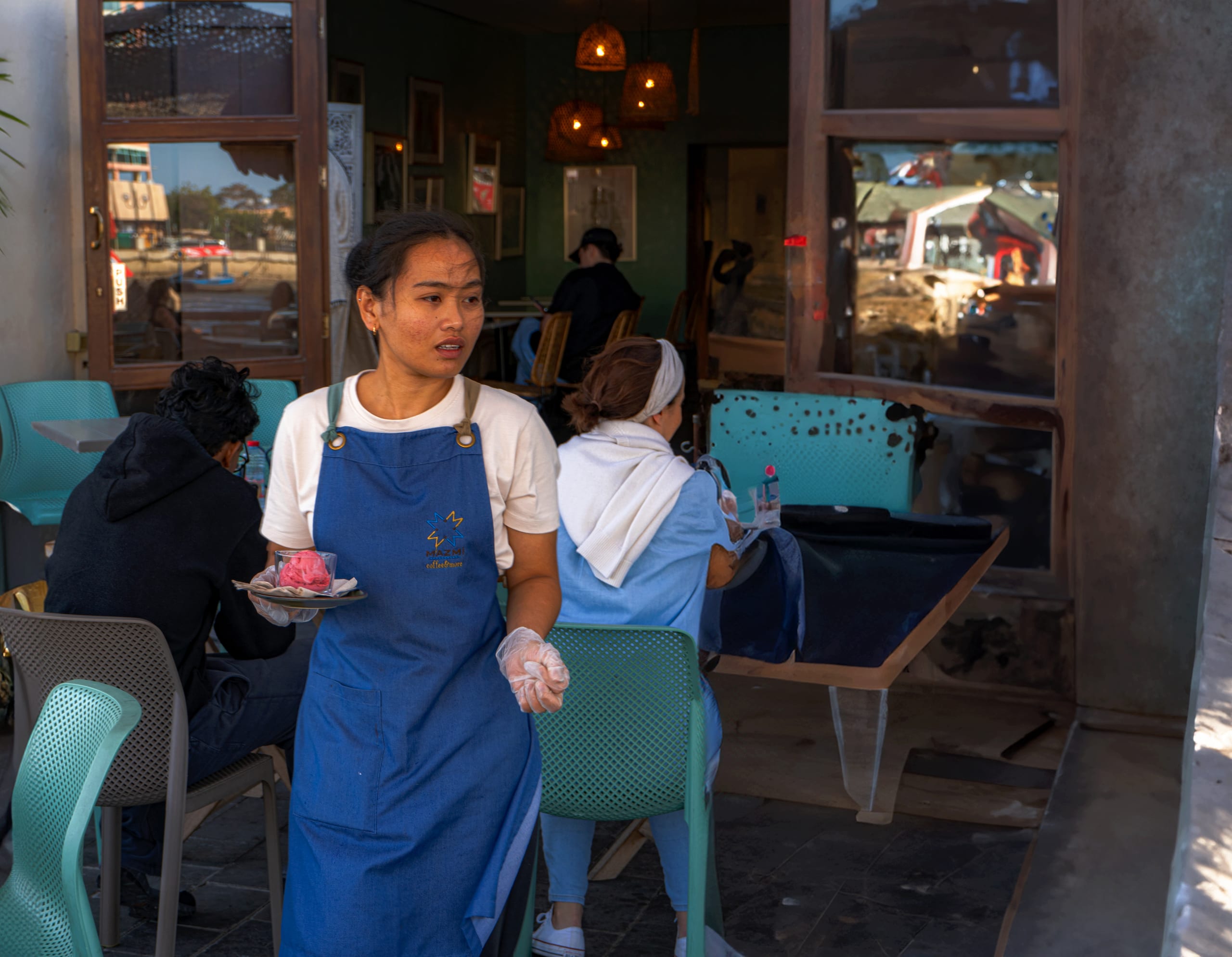 A woman in a blue apron and white t-shirt walks through a restaurant carrying a small plate with a pink dessert, surrounded by seated patrons and turquoise chairs.