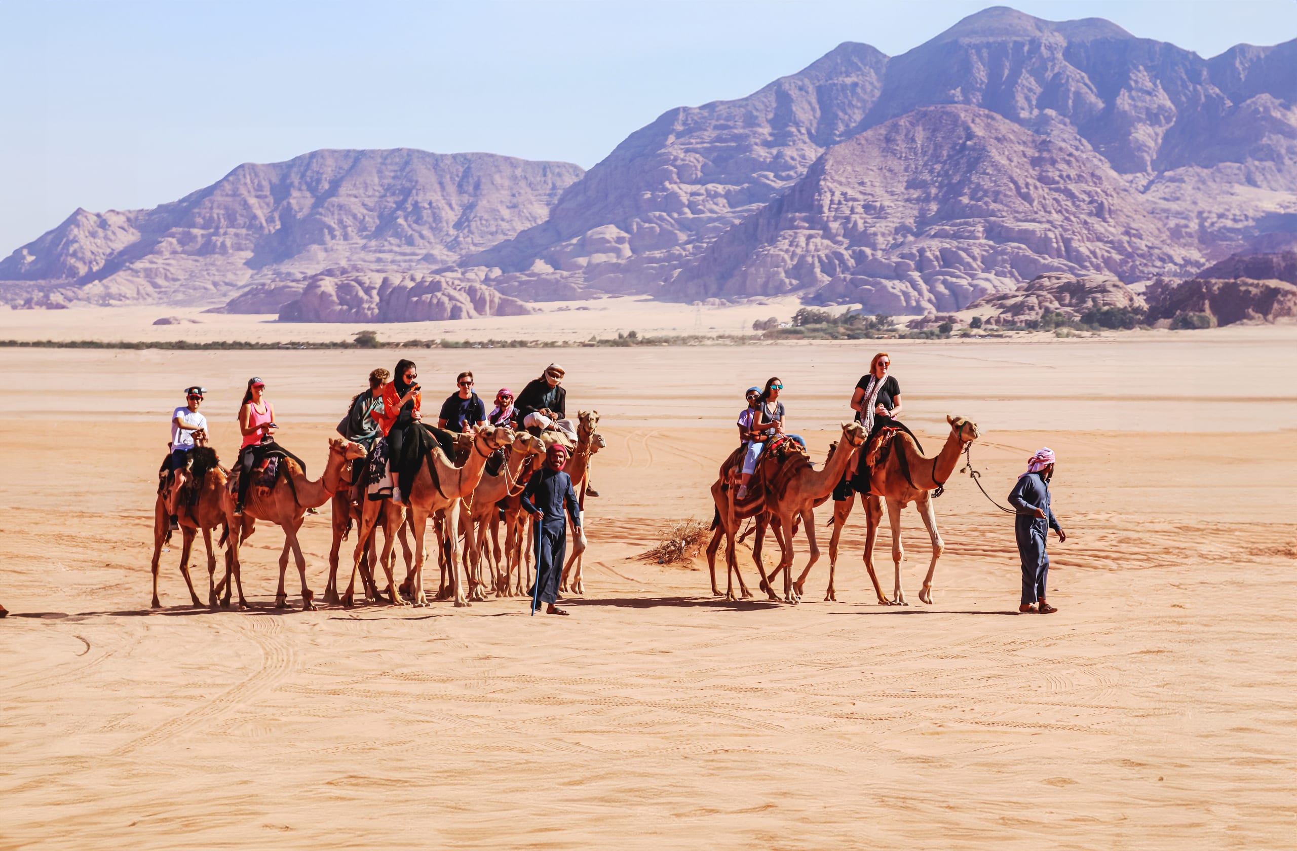 A group of people riding camels in a wide desert landscape with rocky mountains in the background, led by guides walking on the sand.