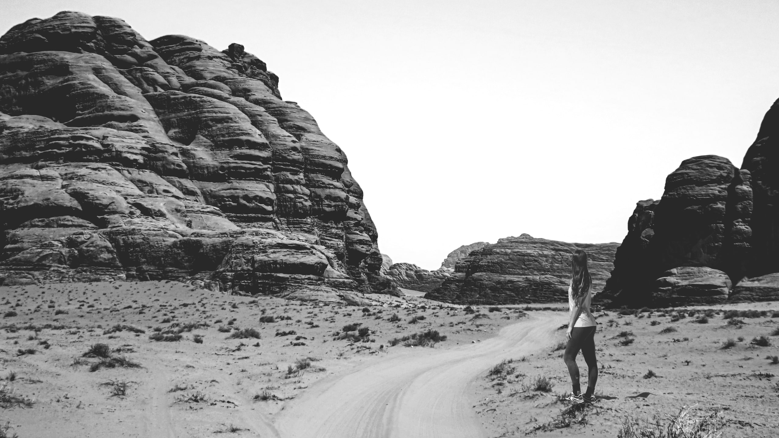 A woman stands on the right side of a wide, sandy desert path, looking toward towering dark rock formations under a bright white sky in a high-contrast monochrome photograph.
