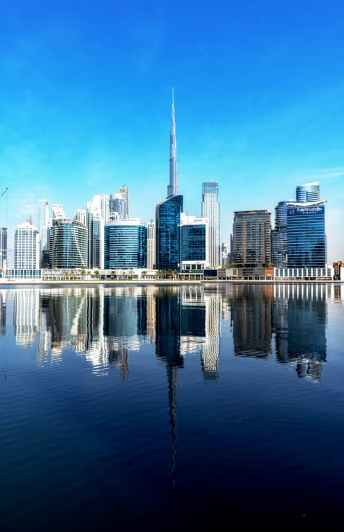 Daytime view of a city skyline featuring a very tall spire building centrally framed, with sharp vertical reflections of the entire row of skyscrapers cast across calm dark blue water.