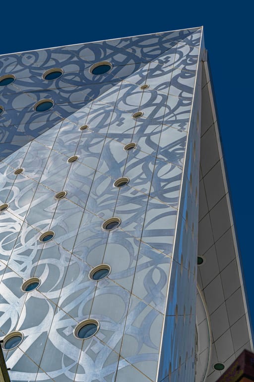 Low-angle view of a modern architectural facade with circular portholes and glass panels covered in large decorative Arabic calligraphy against a deep blue sky.