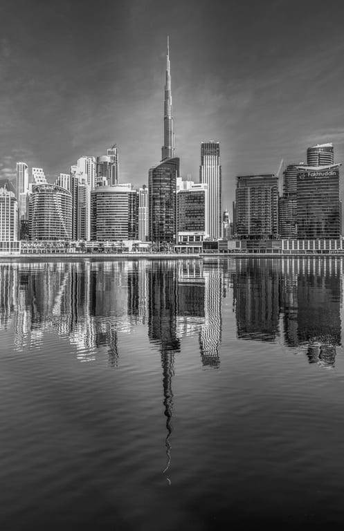 Monochrome photograph of a tall skyscraper towering over a modern city skyline, with its sharp reflection mirrored perfectly on the still water surface in the foreground.