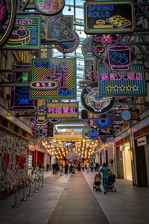 Low-angle shot looking down an indoor arcade crowded with glowing neon signs suspended overhead, with shoppers walking below and traditional lanterns lining the distance.