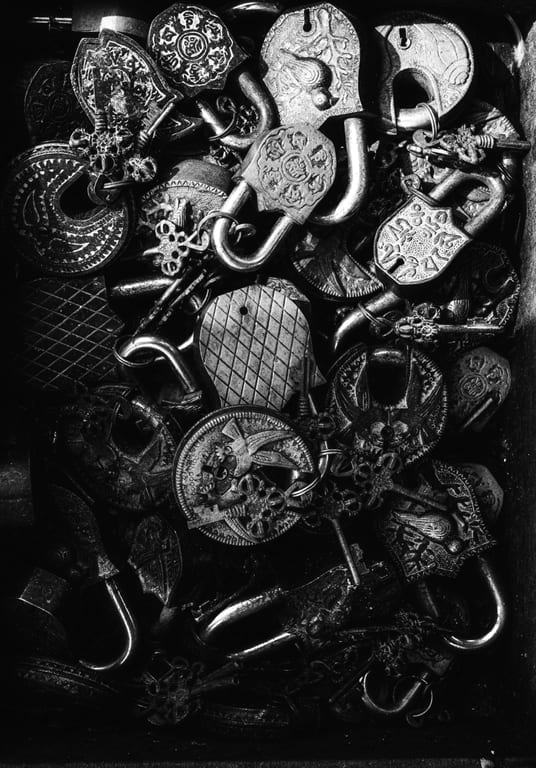 Overhead monochrome close-up of a dense pile of ornate metal padlocks and keys, featuring intricate engravings, metallic highlights, and contrasting deep shadows.