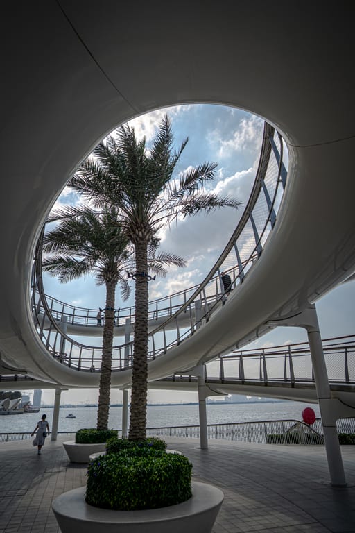 A low-angle view looking upward through a large oval cutout in a white modern architectural structure, revealing two palm trees, a curving elevated walkway with railings, and a body of water in the background.