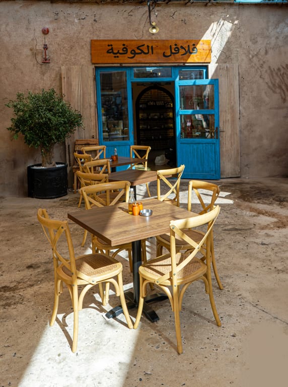 A weathered outdoor courtyard with a square wooden table surrounded by four light wood chairs in the foreground, leading to a small shop with bright blue doors and textured earthen walls.