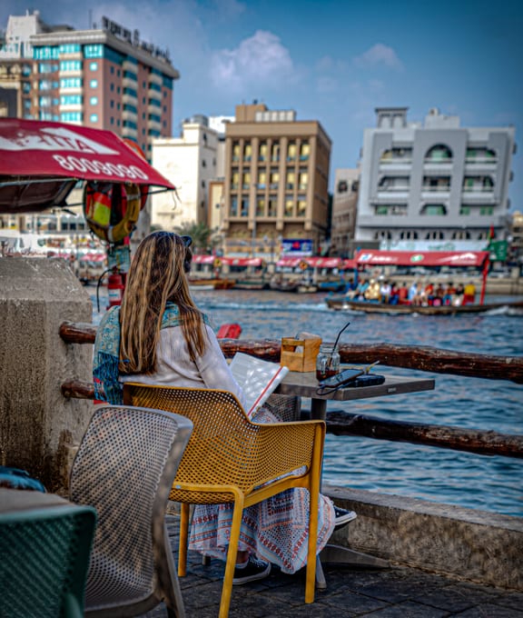 A woman with long blonde hair seen from behind, sitting in a yellow chair at a wooden outdoor table by the water, reading a large open book. Across the water, classic wooden boats carry passengers past a cityscape of traditional and modern buildings under a bright sky.
