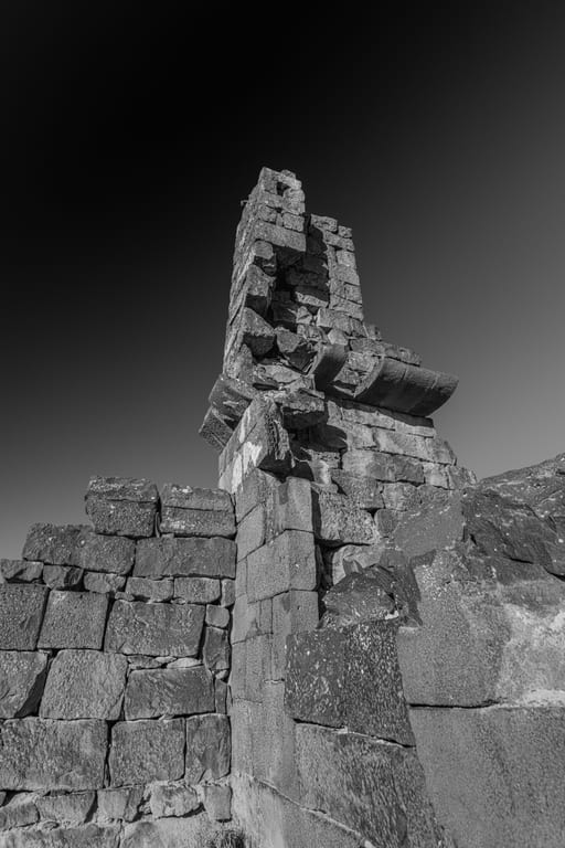 Low-angle black-and-white photograph of ancient stacked stone ruins and a fractured vertical column standing against a gradient sky.