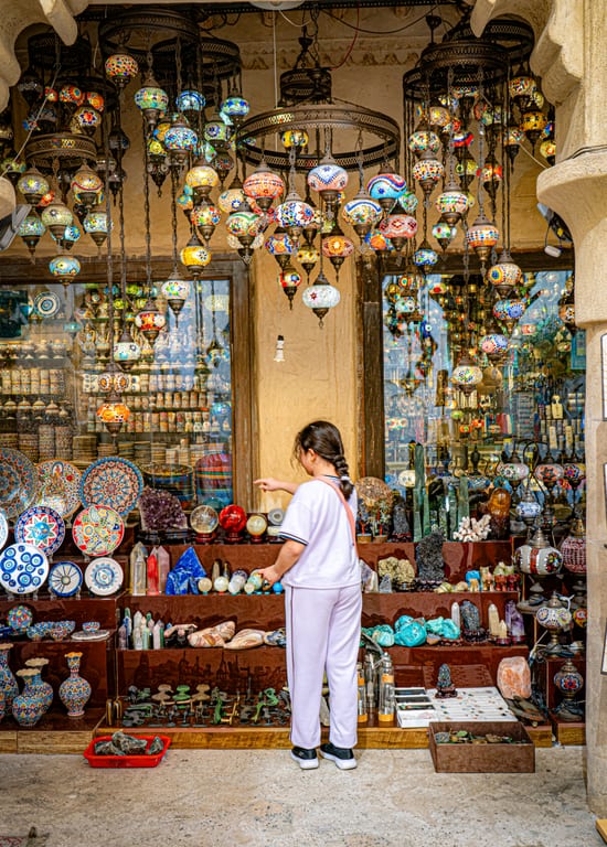 A young person in light-colored casual clothes stands with their back to the camera, reaching toward a crowded display shelf of crystals, minerals, and ceramics inside a shop filled with glowing mosaic glass pendant lamps.