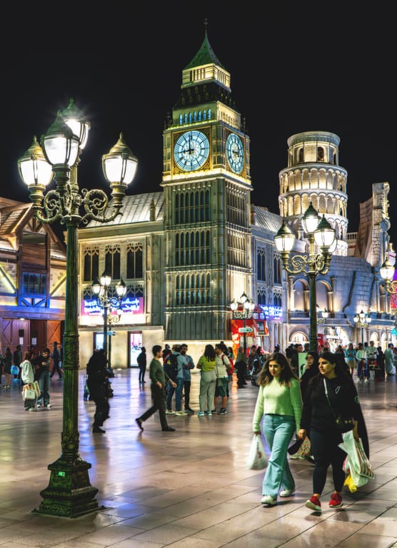 Pedestrians walking on a polished stone plaza at night beneath elaborate traditional streetlamps, with a lit clock tower and classical facades in the background.