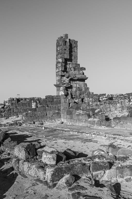 Monochrome low-angle view of a fragmented tall stone column rising above low archaeological stone walls and rubble under a clear sky.
