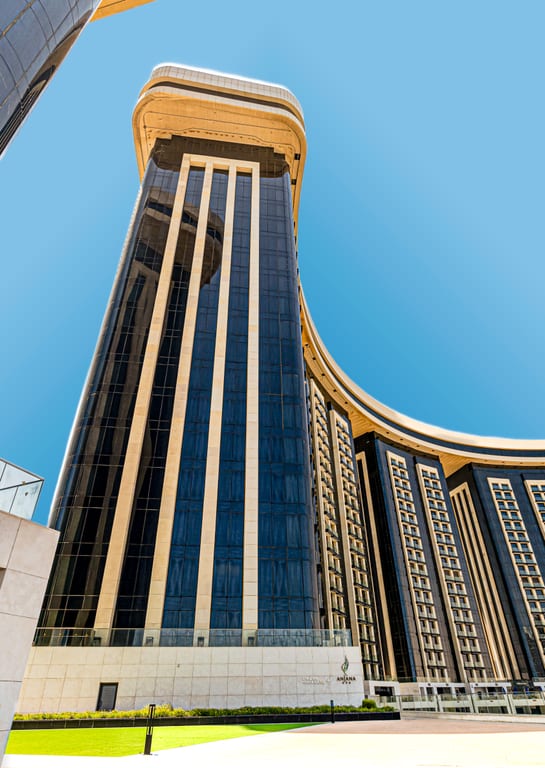 Low-angle upward view of a large modern building featuring dark glass windows, vertical light-colored pillars, and a curving roofline against a clear blue sky.