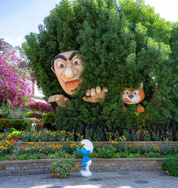 A large sculpted face and hands emerge from a dense green bush alongside a sculpted cat, positioned above a stone planter with yellow and pink flowers where a small blue figurine with a white hat pushes a flower cart.