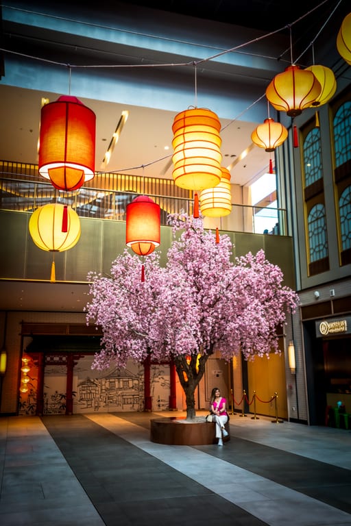 A woman in a pink top sits on a circular wooden bench beneath a large faux cherry blossom tree inside a modern multi-level mall, surrounded by large glowing yellow and red paper lanterns hanging overhead.