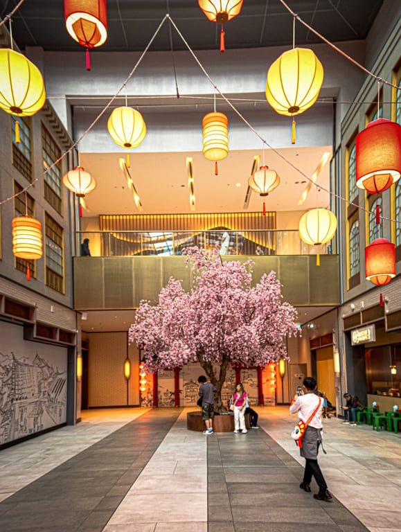 An interior shopping space decorated with dozens of glowing red and yellow paper lanterns suspended on strings across the ceiling above a large blooming pink cherry blossom tree and strolling visitors.