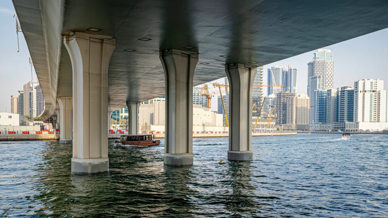 Low-angle photograph looking underneath a large concrete bridge supported by heavy vertical pillars, revealing a waterfront city skyline with modern high-rise towers in the background across calm water.
