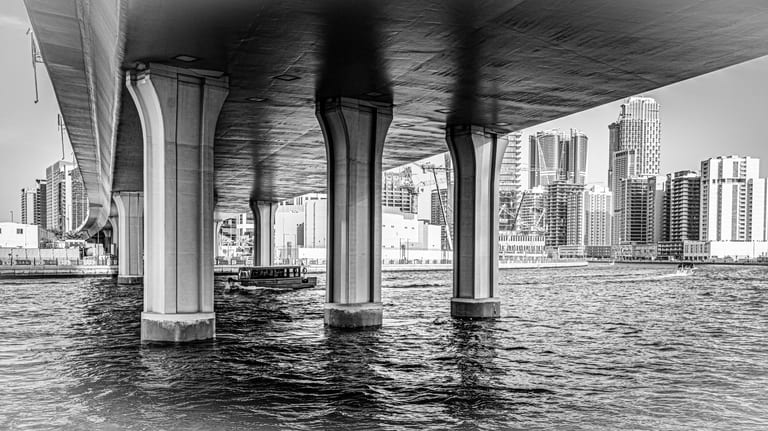 Black-and-white architectural view looking underneath a large highway bridge supported by massive concrete columns planted in dark water, with a small boat crossing beneath and high-rise buildings visible in the background.