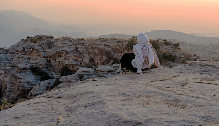 A woman wearing a hijab and floral patterned top sits with her back to the camera on a wide stone cliff edge, looking out over distant layered mountain ridges at sunset.
