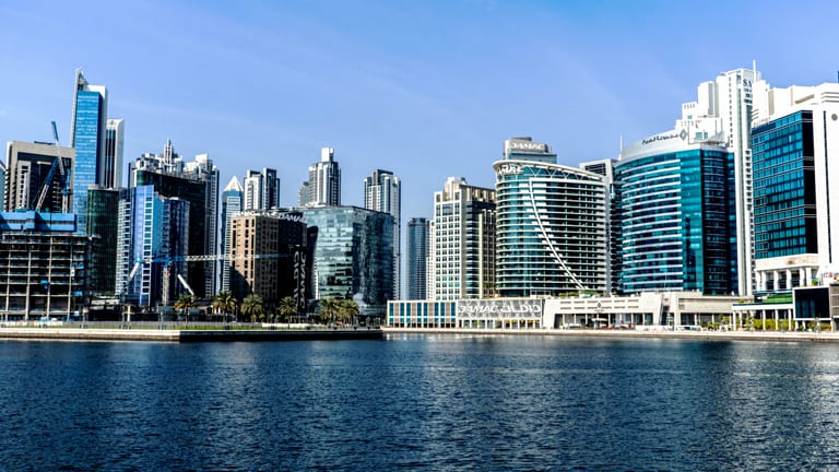 Wide-angle exterior photograph of a modern skyline with numerous skyscrapers along a waterfront, featuring blue glass facades, cranes, and a calm sea in the foreground under a clear sky.