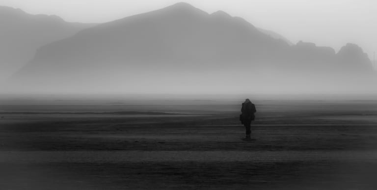 A lone figure walks right in a wide, misty landscape under massive mountain ranges in monochrome.