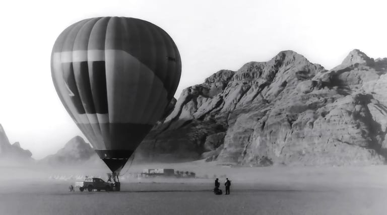 Monochrome photograph of a large hot air balloon grounded on a vast sandy plain, with two small figures standing nearby and towering rocky mountains in the background.