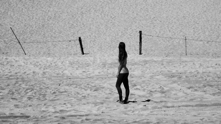 A person with long hair stands in profile on a textured sandy surface, viewed mid-distance with a simple wire fence crossing the upper background in a black-and-white composition.