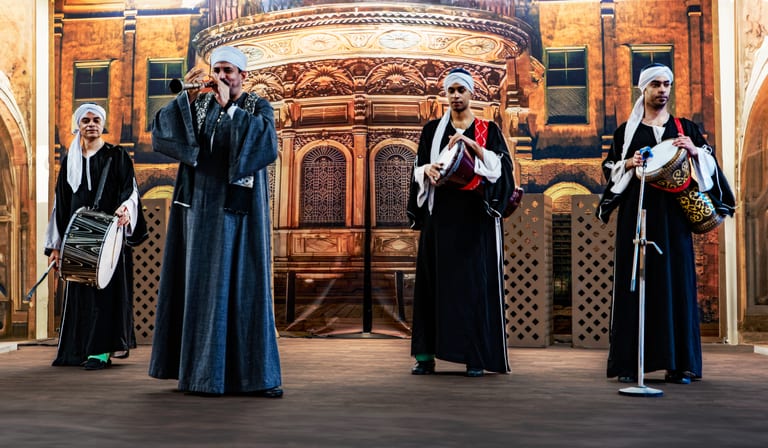 Four male musicians wearing black robes and white turbans stand in a row on a stage, playing drums and a wind instrument, with a large architectural backdrop behind them.