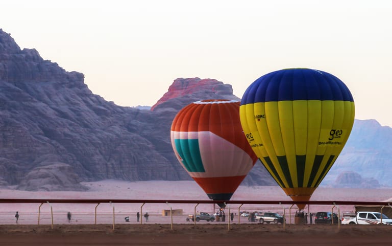 Two hot air balloons, one orange-and-white and one yellow-and-blue, stand grounded in a desert landscape with rocky mountains in the background and a horizontal railing in the foreground.