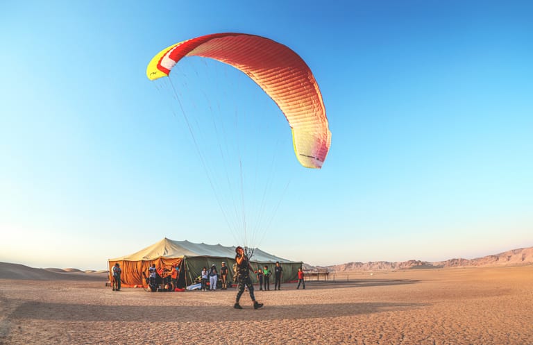 A person standing in a desert landscape holds the suspension lines of a large red and white paraglider canopy, with a large tent and several figures gathered in the background under a blue sky.