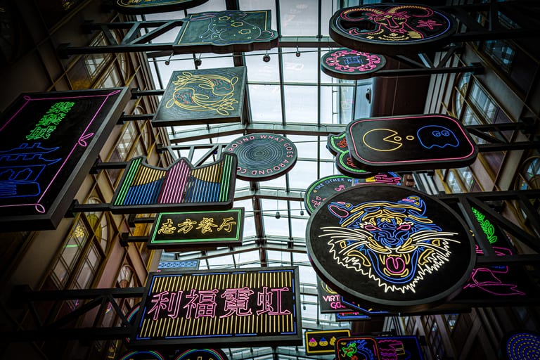 Low-angle view of numerous colorful neon signs hanging from a dark metal grid framework beneath a bright glass atrium roof.