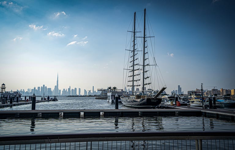 A large two-masted traditional sailing ship moored at a wooden and concrete marina pier, with a city skyline visible across the water in the hazy background under a bright sky.