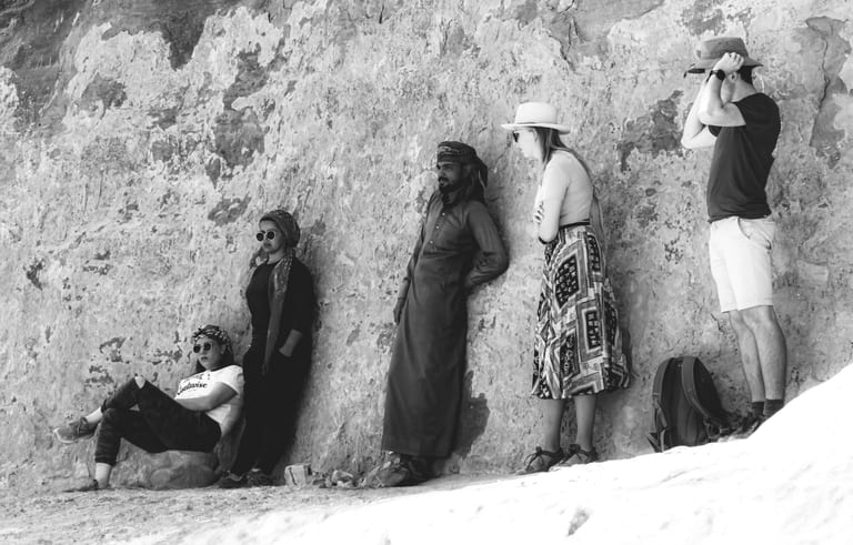 A black and white photograph of five people lined up leaning against a rough stone cliff face, varying in posture from sitting on the ground to standing.