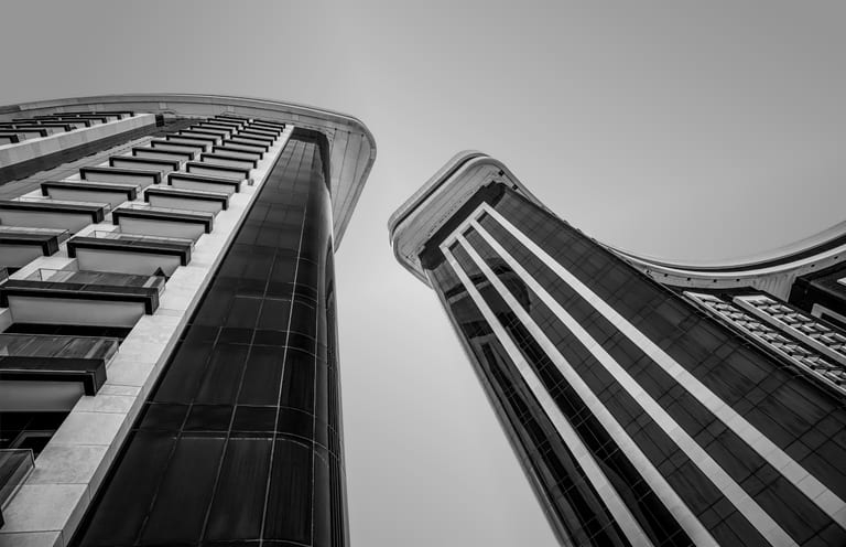 A low-angle black-and-white architectural photograph looking straight up at two modern curved skyscrapers with dark glass windows and contrasting white structural ribs, separated by a slice of pale sky.
