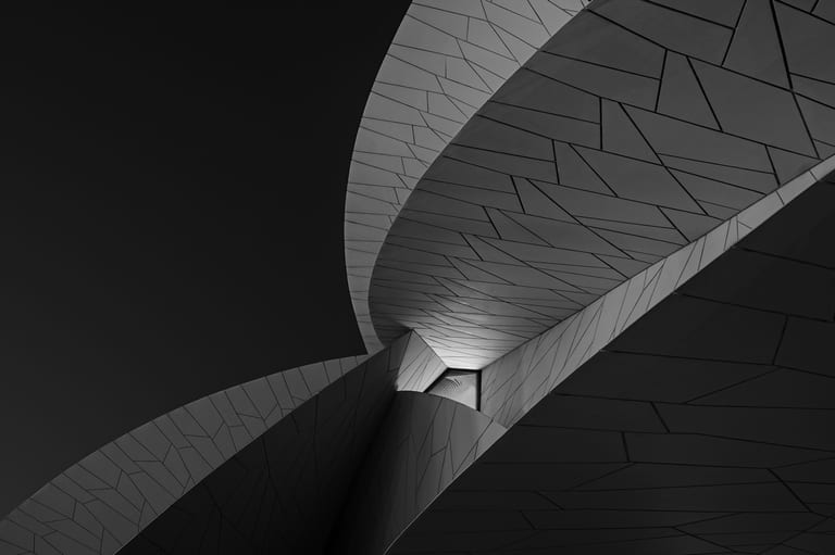 A low-angle black and white photograph of interlocking curved architectural panels forming abstract geometric shapes against a dark sky, illuminated by light from within a central recess.