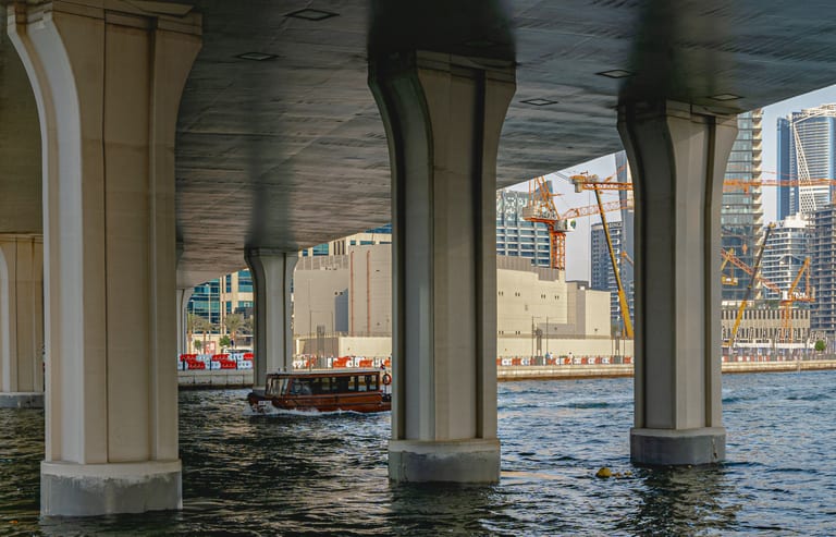 A low-angle exterior shot looking across open water beneath a large concrete bridge supported by heavy pillars, with a small wooden boat moving across the midground and urban skyscrapers in the distant background.