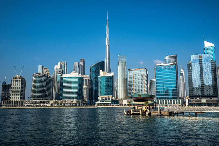 Wide daylight view across a body of water toward a dense city skyline, dominated by a towering skyscraper at the center under a clear blue sky.