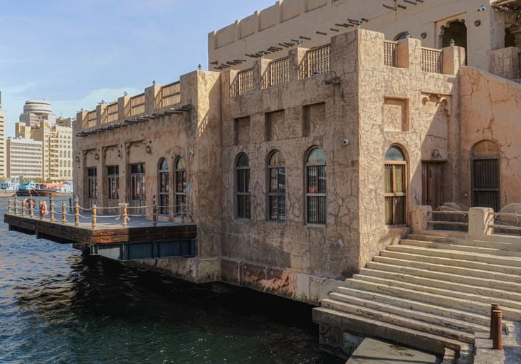 Exterior view of traditional low-rise stone and concrete waterfront architecture with arched windows, wooden balconies, and a set of exterior stone stairs leading down to dark water, with modern skyscrapers visible in the distant background under a bright sky.