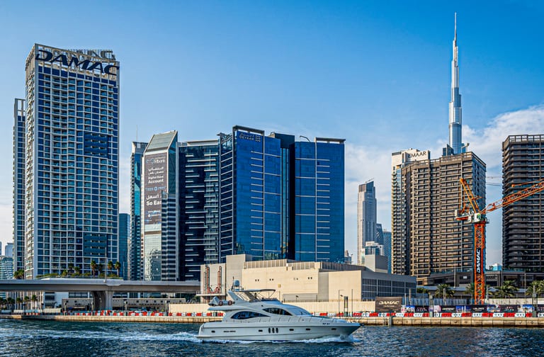A white yacht moving across a blue canal water in front of a dense urban skyline featuring tall skyscrapers, modern buildings, and a bright clear sky.