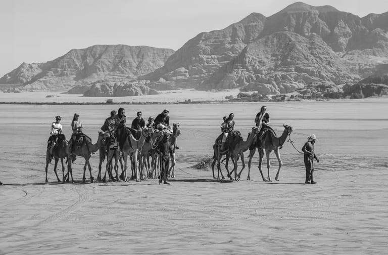 Monochrome wide shot of a caravan of people riding camels across a bright desert floor with layered mountain ranges in the background.