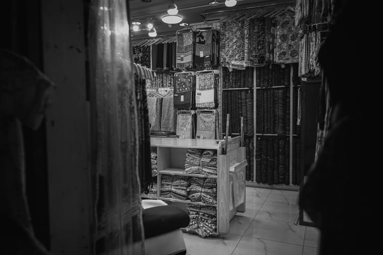 Monochrome interior view of a textile shop, featuring neatly folded stacks of fabric on wooden shelving and numerous hanging scarves under bright overhead lights, framed by dark silhouettes in the foreground.