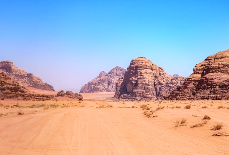 Wide-angle desert landscape view showing sandy foreground tracks, sparse dry shrubs, towering red-brown sandstone rock formations in the midground, and a pale blue sky stretching overhead.