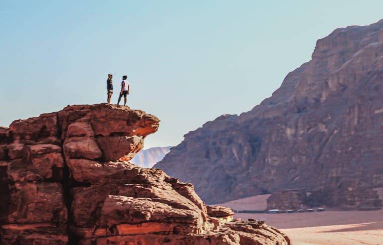 Two people standing on the rocky outcrop of a desert cliff, viewed against a vast expanse of mountains and clear sky.