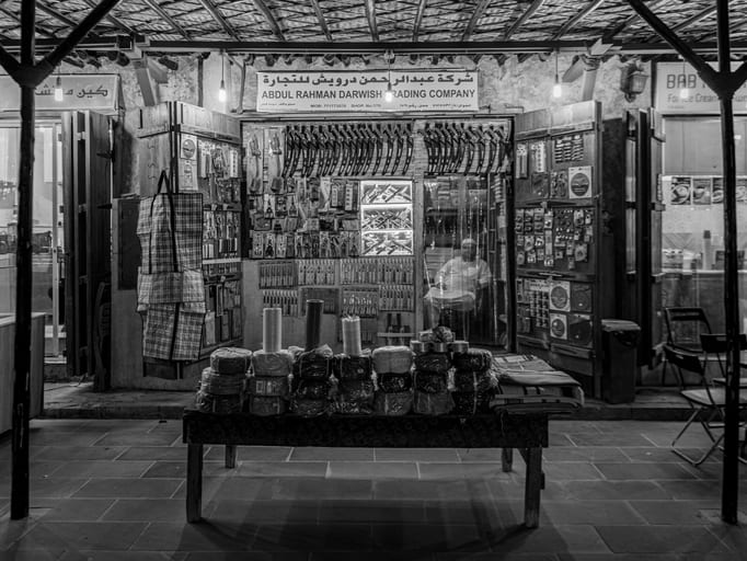 A black-and-white documentary photograph of a storefront featuring walls densely lined with displayed tools and hardware. A wooden table in the foreground holds stacked bundles of goods, and a seated shopkeeper is visible inside the dim interior.
