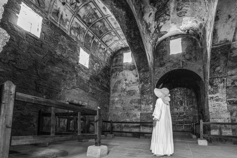 A full-length monochrome photograph of a figure in a white dress and wide-brimmed hat standing inside a vaulted stone hall with ancient frescoes, looking upward toward murals and high rectangular windows.